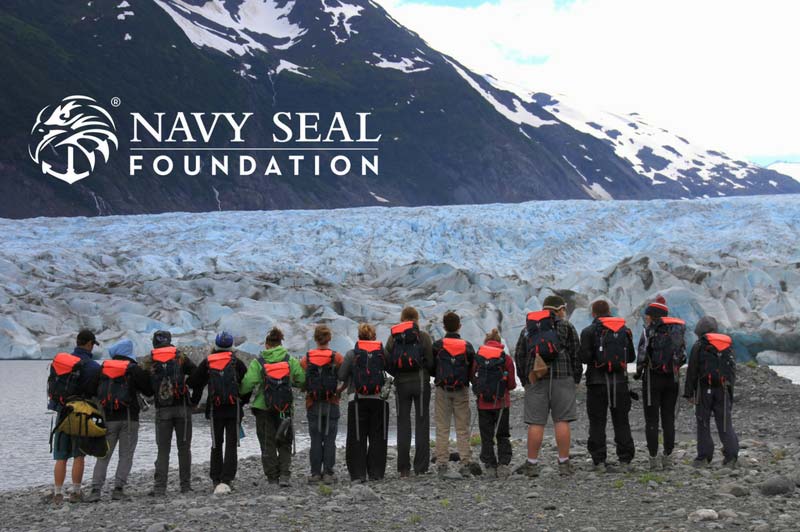 People with backpacks stand by a glacier, with "Navy Seal Foundation" text. North to the future is appropriate.
