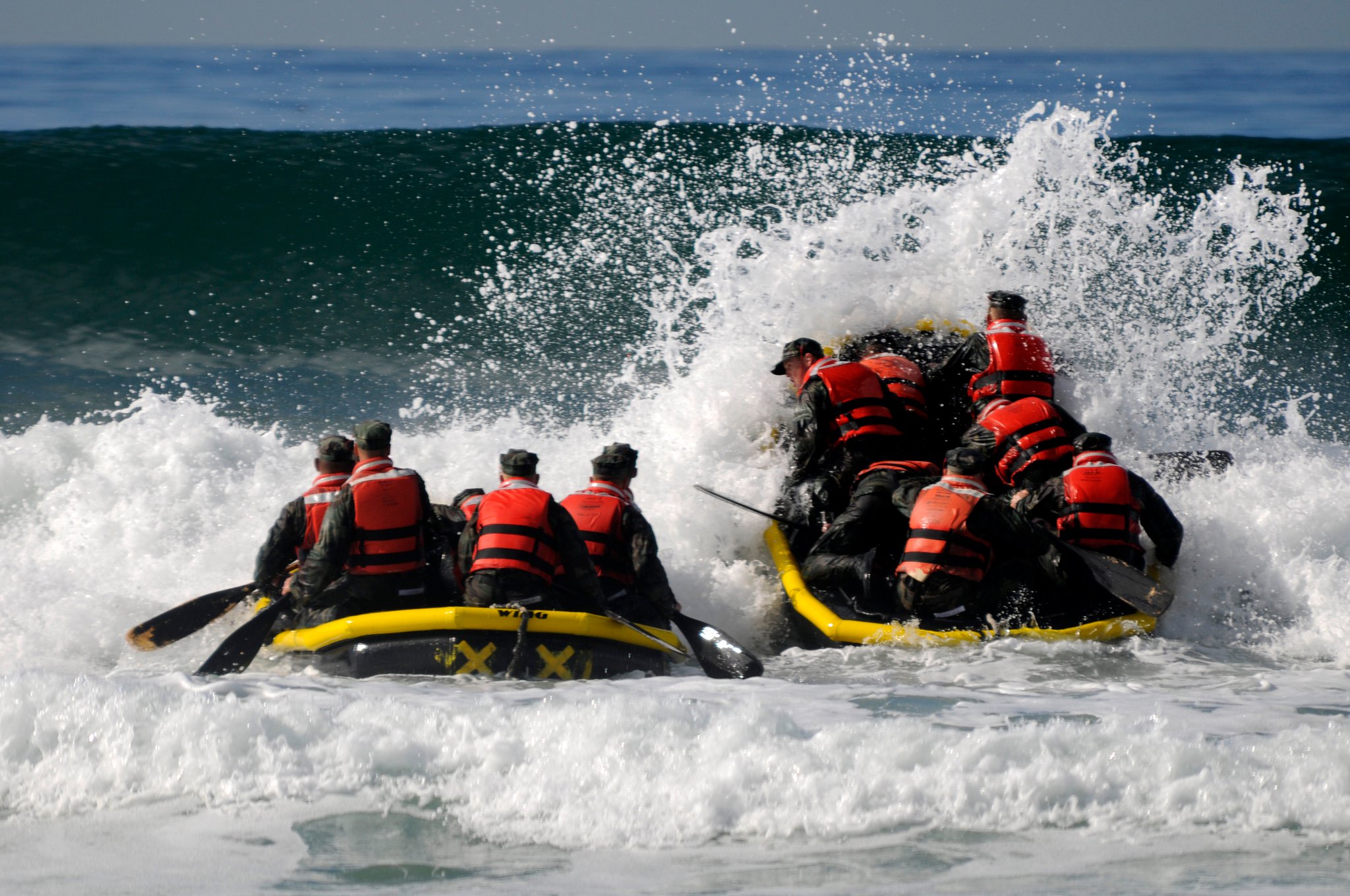 Group of Navy SEAL trainees paddling a yellow inflatable boat through rough ocean waves during training, wearing black wetsuits and red life vests.