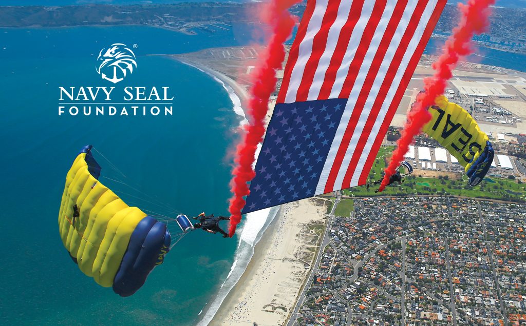 Skydivers with U.S. flag parachutes trailing red smoke, over a coastal cityscape, related to the Navy Seal Foundation.