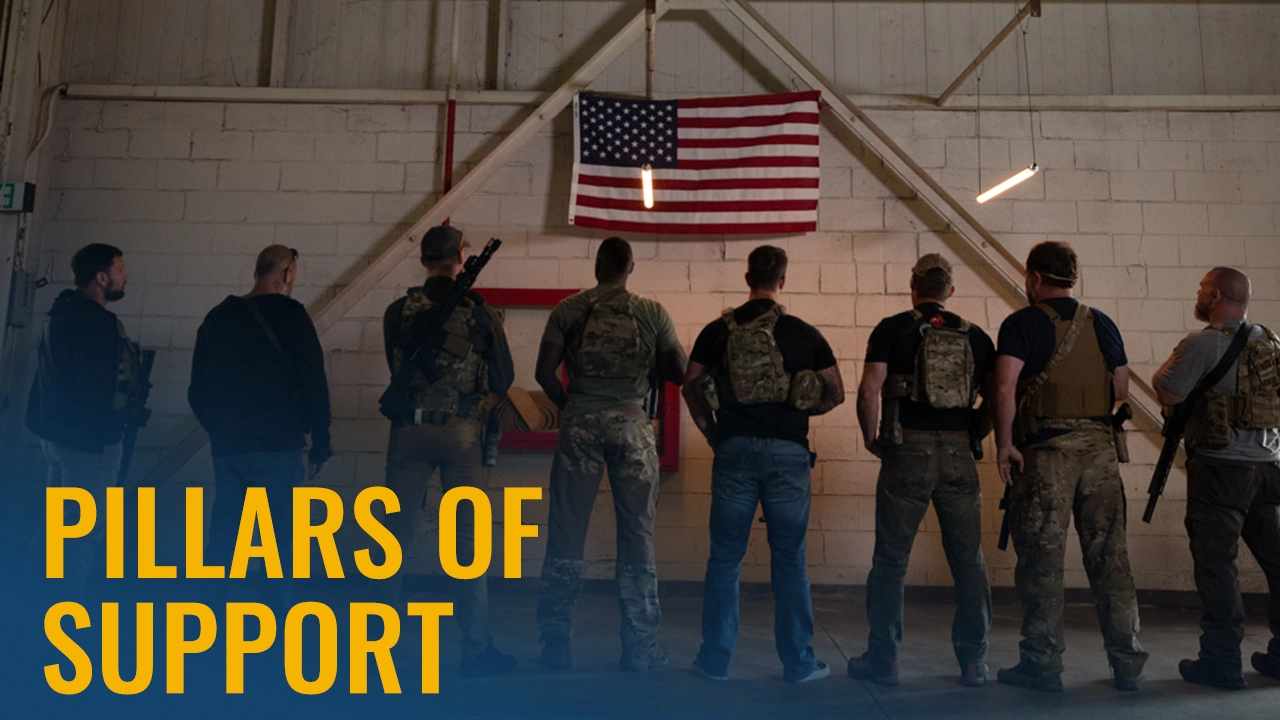 Seven men in tactical, military-style clothing stand with their backs to the camera, facing an American flag hanging on a wall inside an industrial-looking building. The text "PILLARS OF SUPPORT" appears in bold yellow letters at the bottom left of the image.